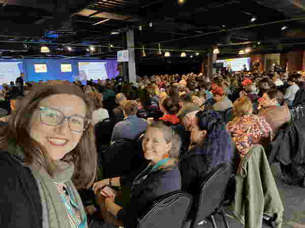 Foto: Vielen Menschen sitzen im Publikum. Im Vordergrund schauen zwei Frauen froh in die Kamera.