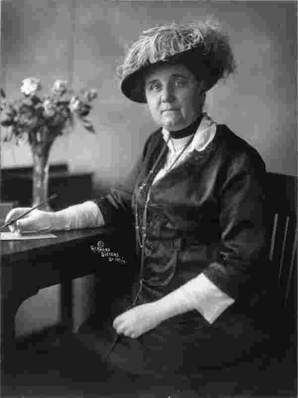 Historisches Fotos: Eine nobel gekleidete Frau sitz an einem Schreibtisch und schaut in die Kamera.