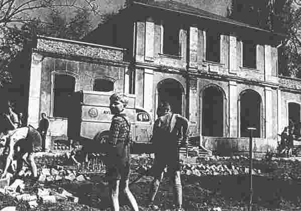 Schwarzweiß Foto: Kinder und Erwachsene stehen von Backsteinen um geben vor einem halten Haus an dem sie bauen.