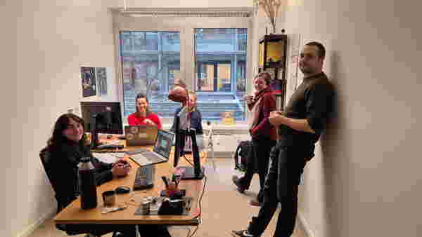 Farbfoto im Querformat: Fünf Personen stehen und sitzen um und an zwei Tischen in einem kleinen Büro Zimmer. Sie wirken in den Moment der Arbeit vertieft.