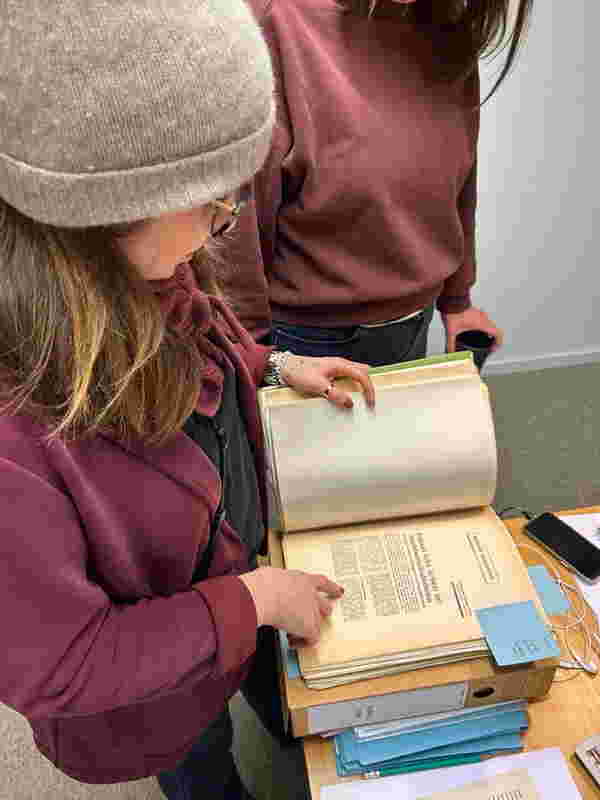 Farbfoto im Hochformat: Zwei Frauen beugen sich an einem Tisch stehend über historische Texte und Ordner. Die Kamera guckt ihn über die Schulter.