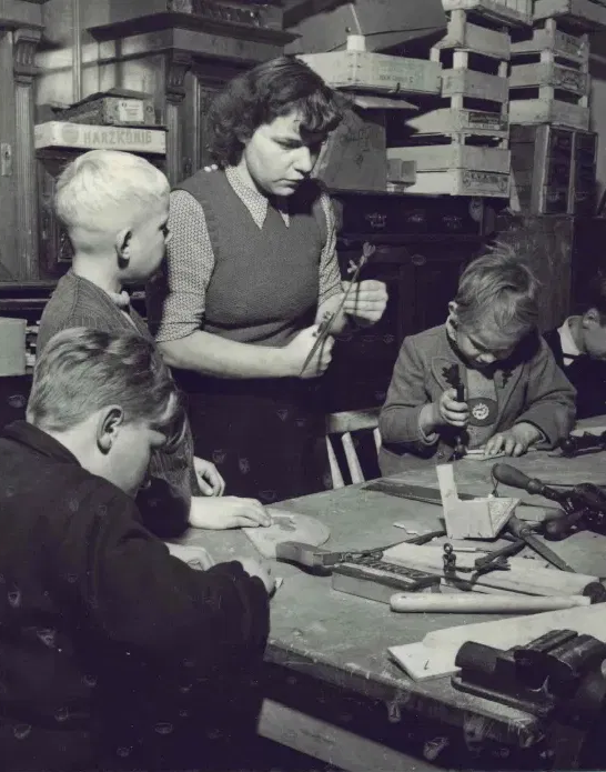 Schwarzweiß Foto einer Werkstatt: Ein Frau leitet drei Kinder bei der Handarbeit an. Sie stehen und sitzen um einen Tisch mit Werkzeug in der Hand.