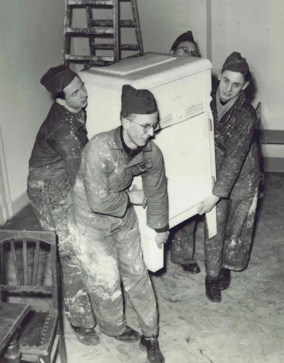 Schwarzweiß Foto: Vier uniformierte Männer tragen eine Ofen aus einem kargen Raum heraus. Im Hintergrund steht eine Leiter.