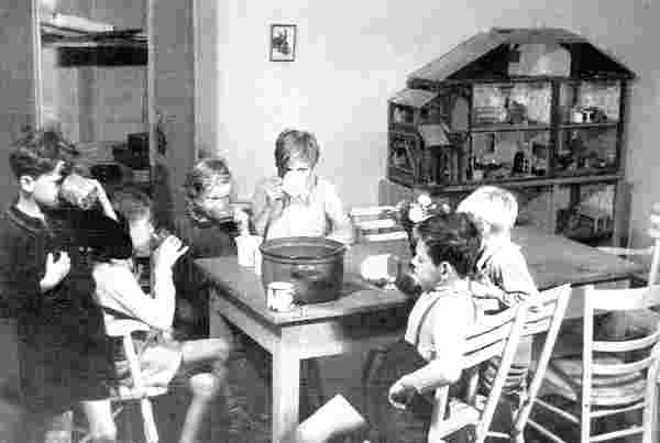 Schwarzweiß Foto im Querformat: Sechs Kinder stehen und sitzen um einen Tische herum und trinken aus Bechern. Auf dem Tisch steht weitere Becher und ein großer Topf. Im Hintergrund steht auf einem Regal ein großes altes Puppenhaus.